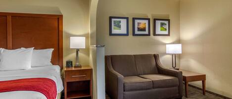 Habitación de hotel con cama, sofá marrón, dos mesitas de noche con lámparas y tres cuadros en la pared.