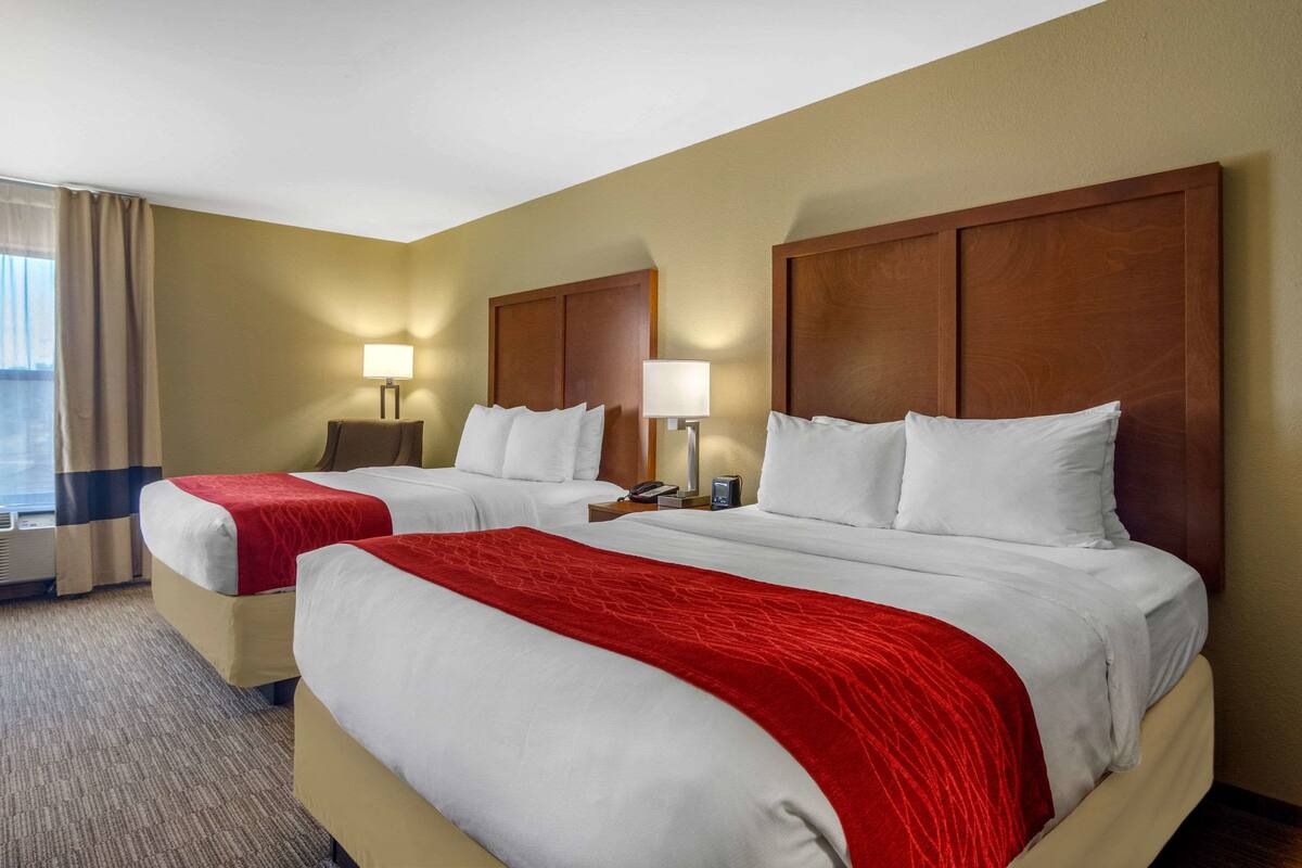 A hotel room with two beds, each with white linens and red throw blankets. There are bedside tables with lamps and a phone. The room has beige walls and a carpeted floor.