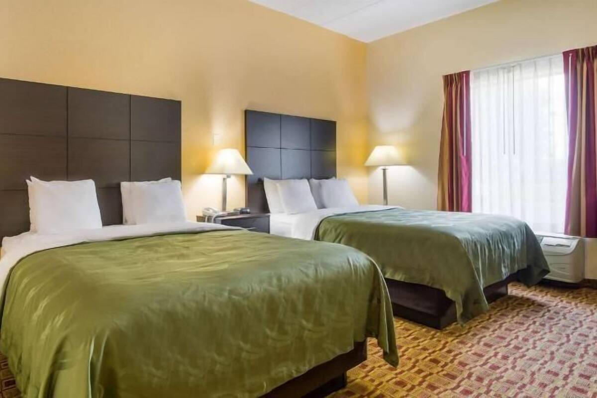 A hotel room with two beds, a nightstand, a lamp, and a window with curtains.