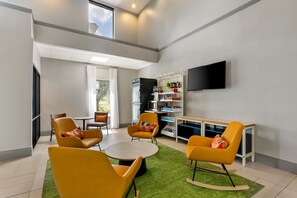 Un salon moderne avec des fauteuils jaunes, une table ronde, un téléviseur à écran plat et un coin cuisine intégré.