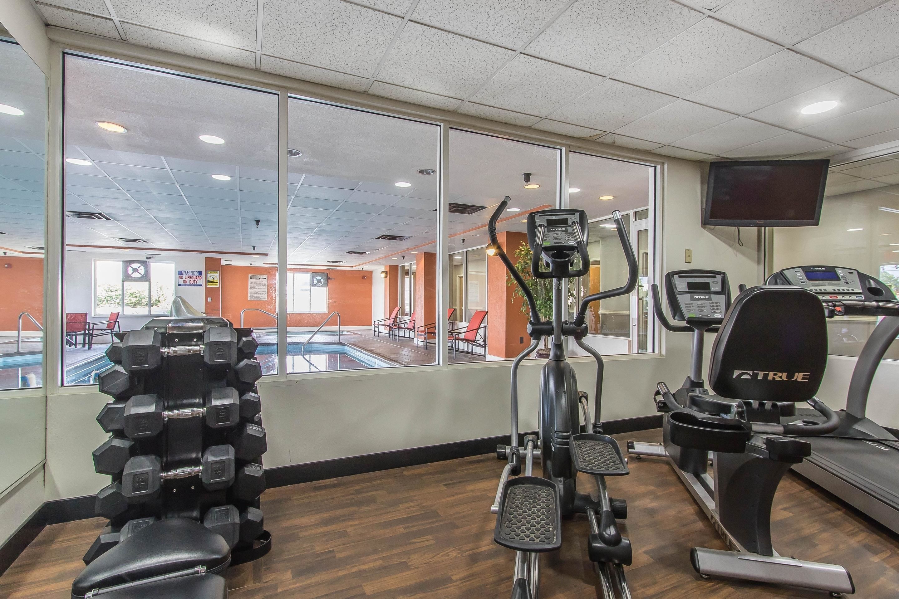 Ett gym med träningsutrustning, stora fönster och ett poolområde som syns genom fönstren.