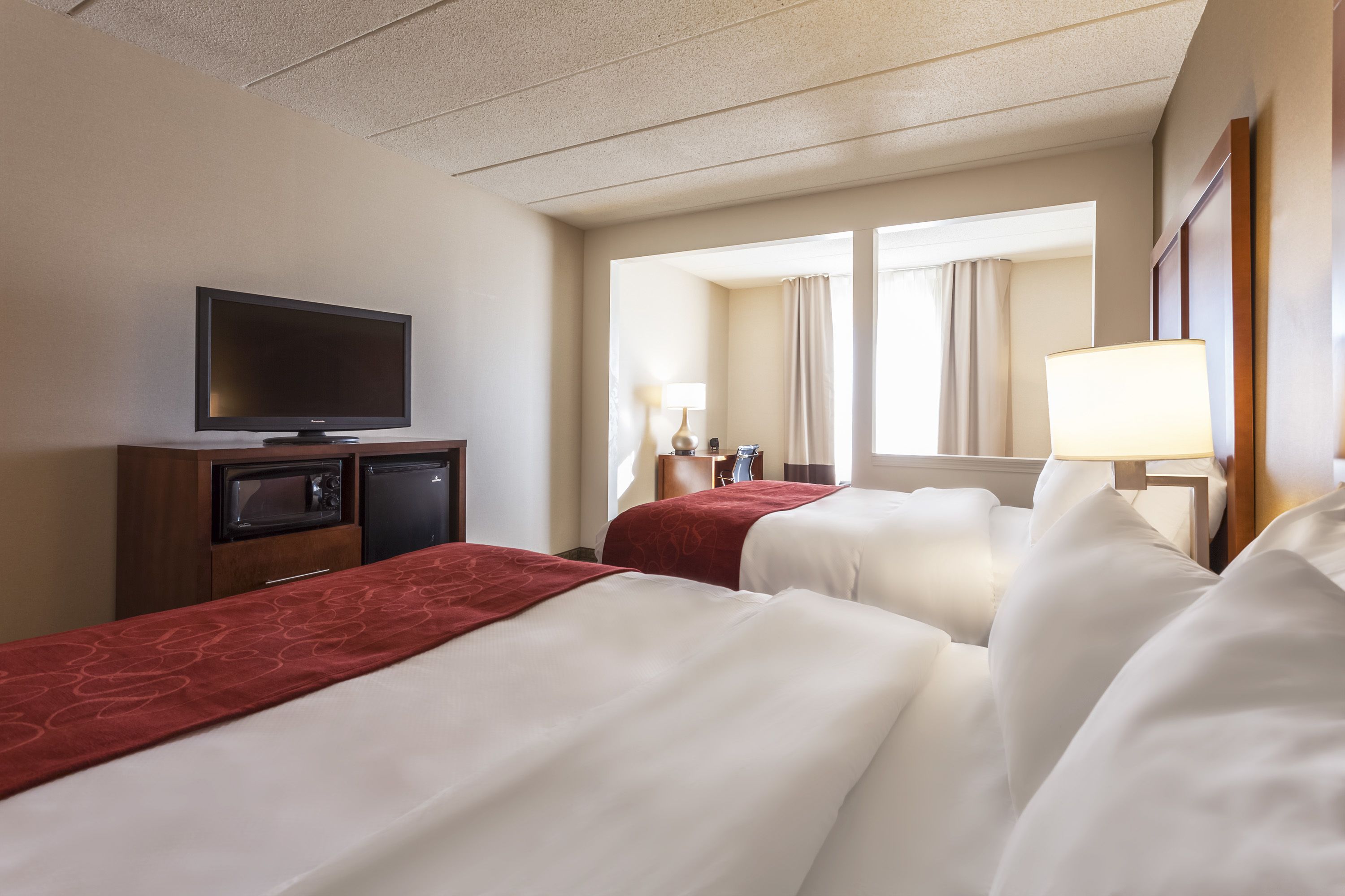 A hotel room with two beds, a flat-screen TV, a minibar, and a window with curtains.