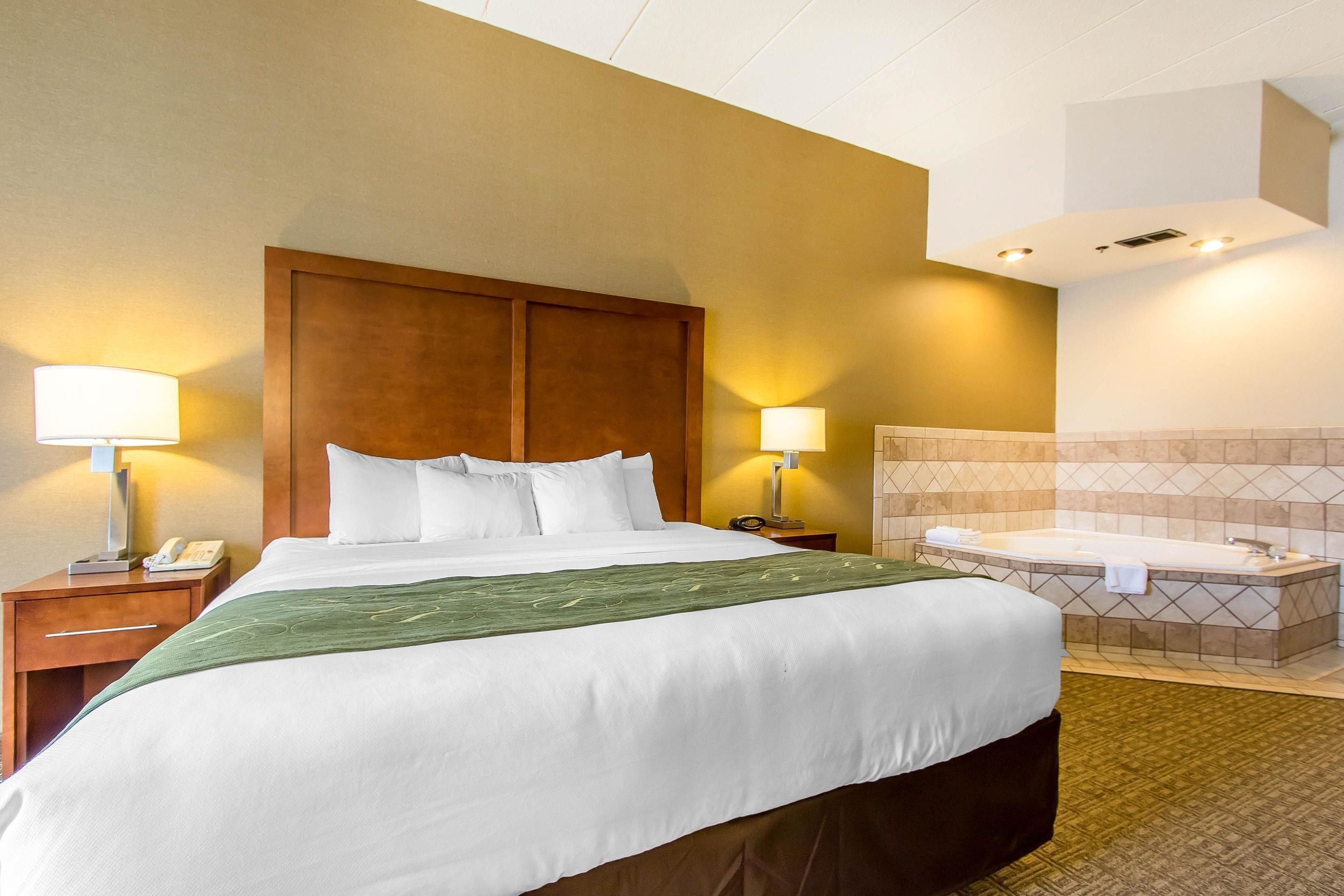 A hotel room with a large bed, two bedside lamps, a wooden headboard, a bathroom with a bathtub, and a tiled wall.