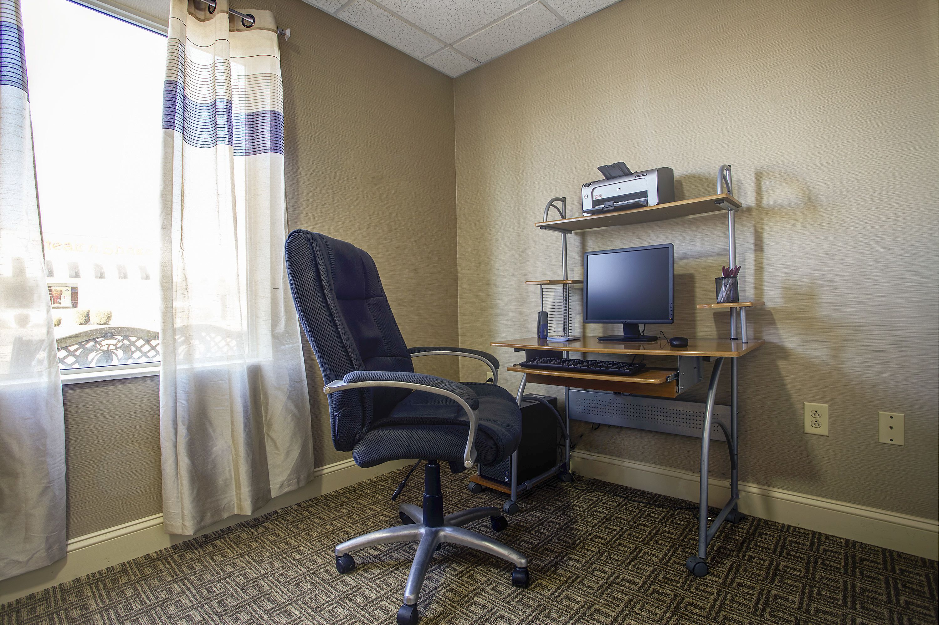 A hotel room with a desk, computer, chair, and a window with curtains.