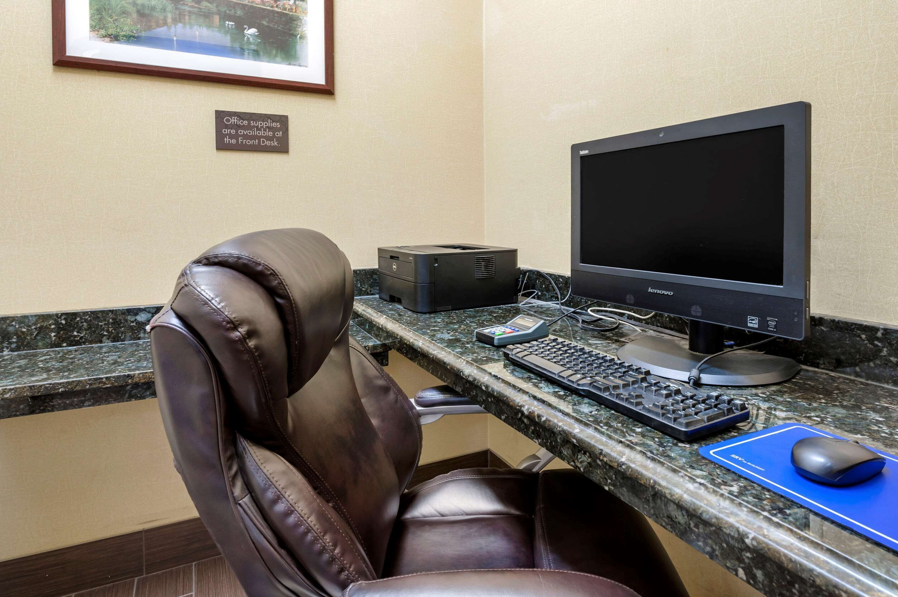 Una oficina de hotel con una butaca de cuero, una computadora de escritorio, una impresora y un cuadro en la pared.