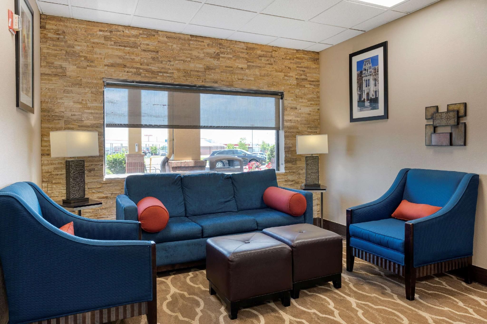 A hotel lobby with a stone wall, a large window, a blue sofa, two blue armchairs, a brown ottoman, and a framed picture on the wall.