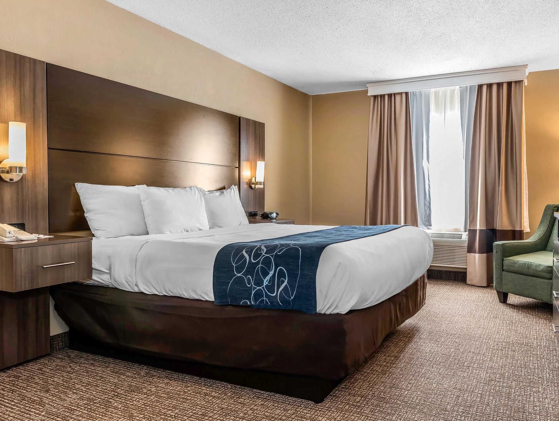 Quarto de hotel com uma cama grande, mesas de cabeceira, escrivaninha, uma cadeira e janela com cortinas.