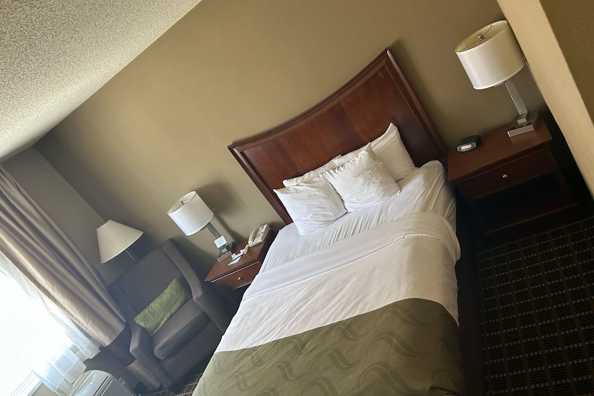 Habitación de hotel con una cama grande, dos lámparas de noche, una mesita de noche con teléfono y lámpara, una ventana con cortinas y un equipo de aire acondicionado.