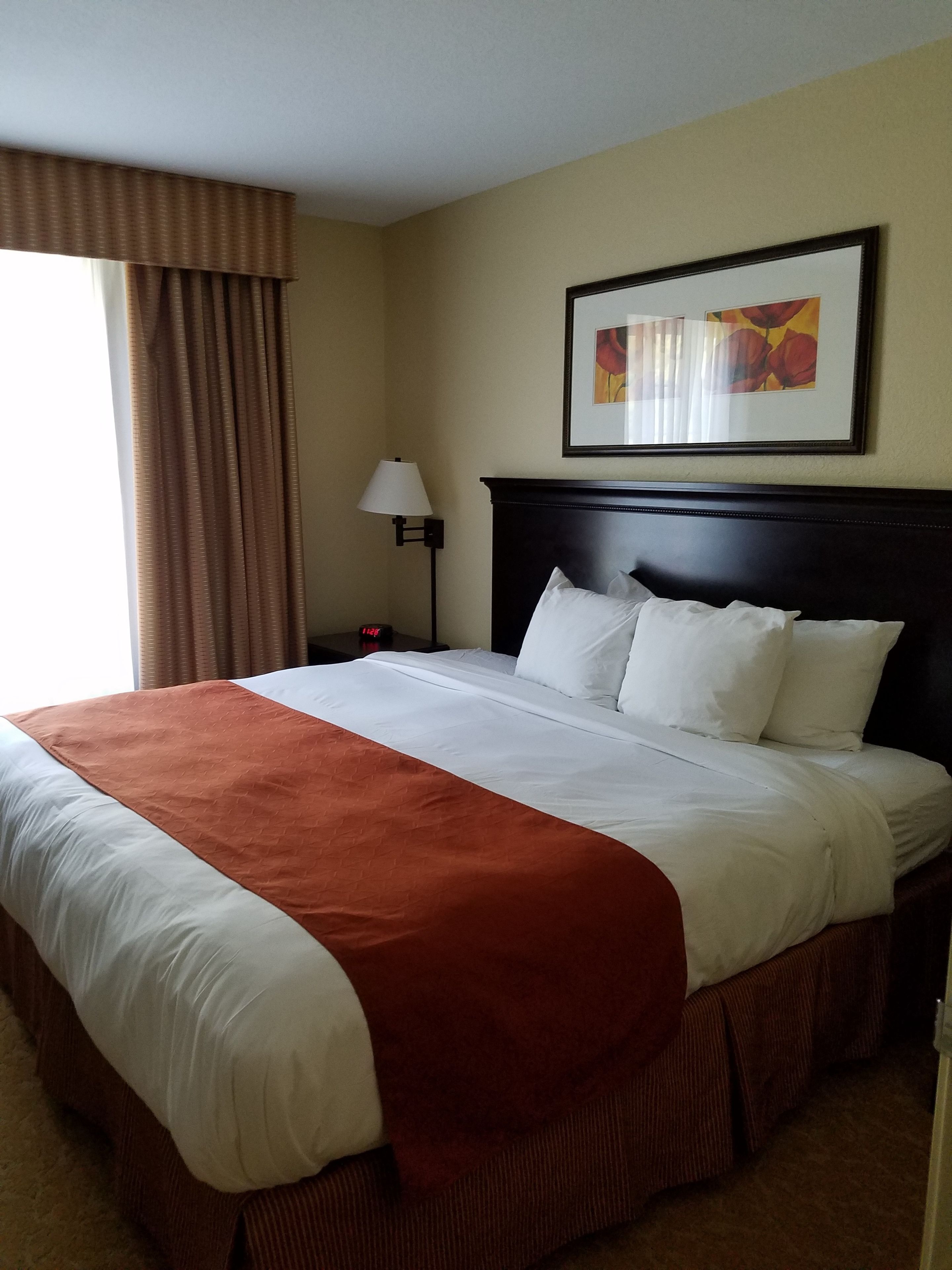 A hotel room with a large bed, a framed picture on the wall, a nightstand with a lamp, and a window with curtains.