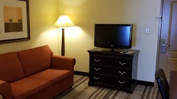 A hotel room with a sofa, a TV on a dresser, a lamp, and a door.