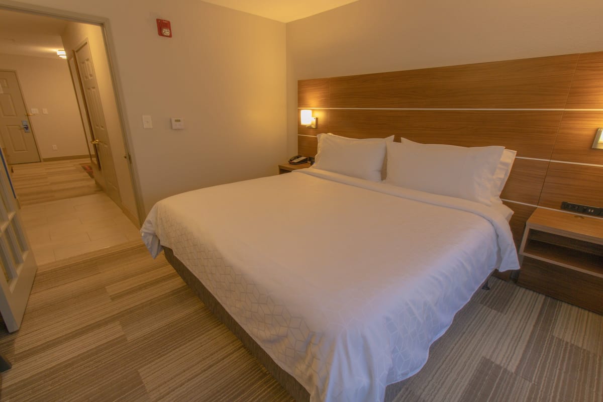 Hotellihuone, jossa on suuri sänky, puinen pääty, yöpöytä lampun kanssa ja matolla peitetty lattia.