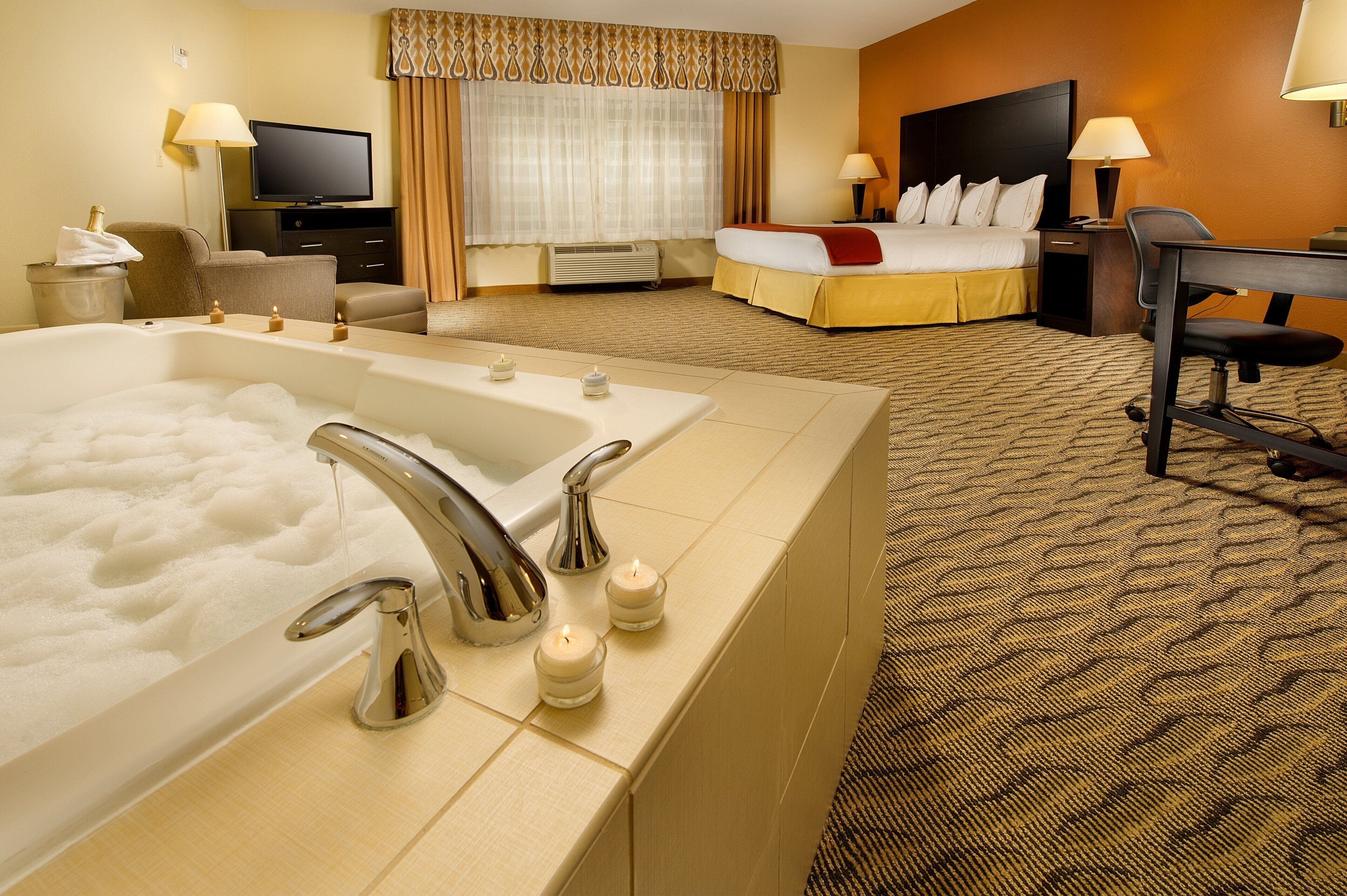 A hotel room with a large bathtub, a bed, a desk, and a television.
