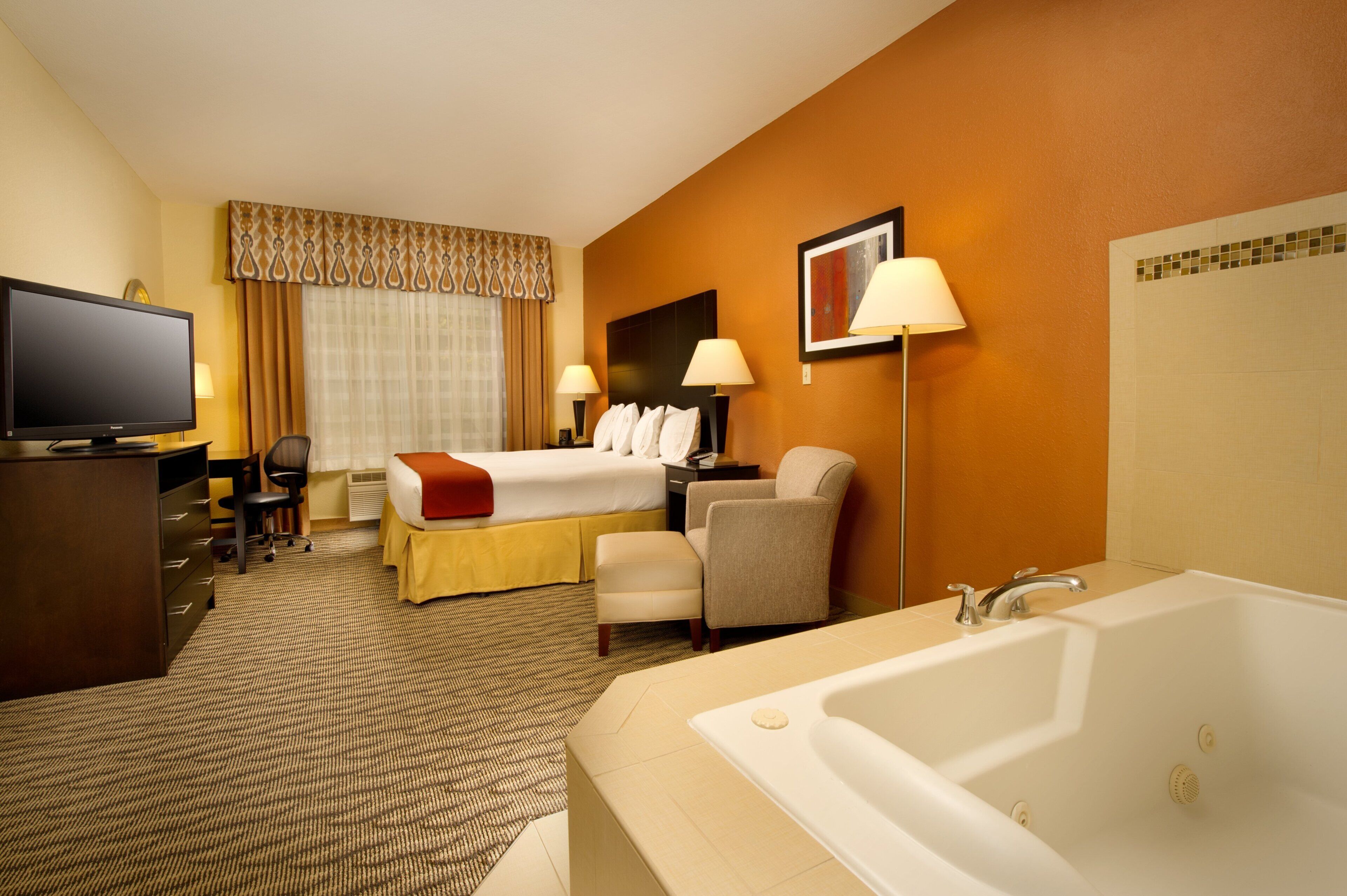 A hotel room with a bed, a desk, a chair, a television, a lamp, and a bathtub.