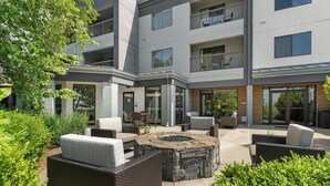 A modern outdoor patio with wicker furniture, a stone fire pit, and a view of a multi-story building with balconies. - Courtyard by Marriott Burlington Williston (Williston)