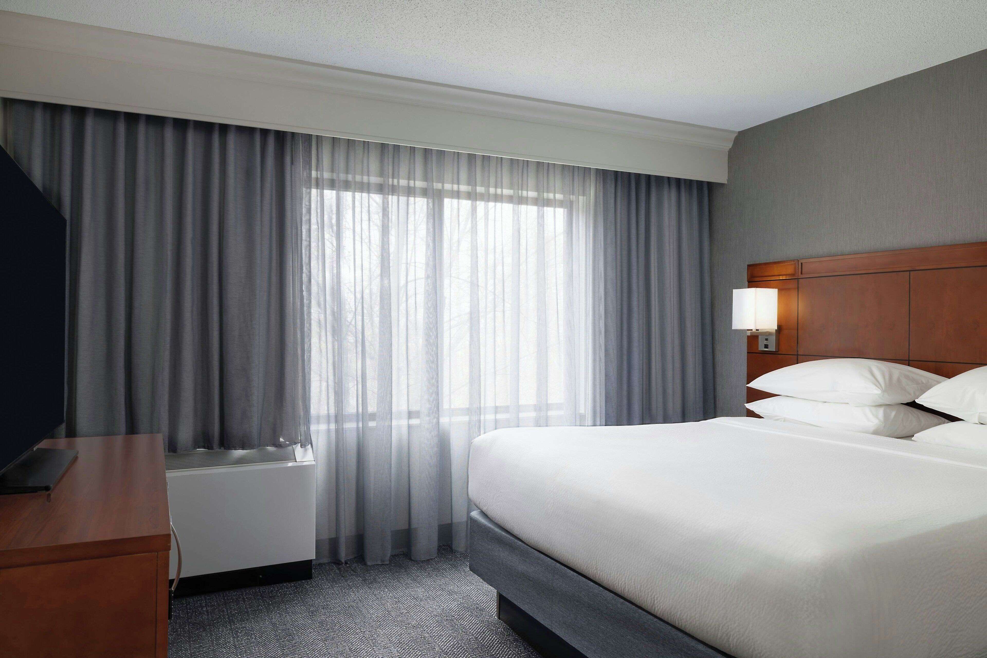 A hotel room with a bed, a desk, a TV, a window with curtains, and a wall-mounted lamp.