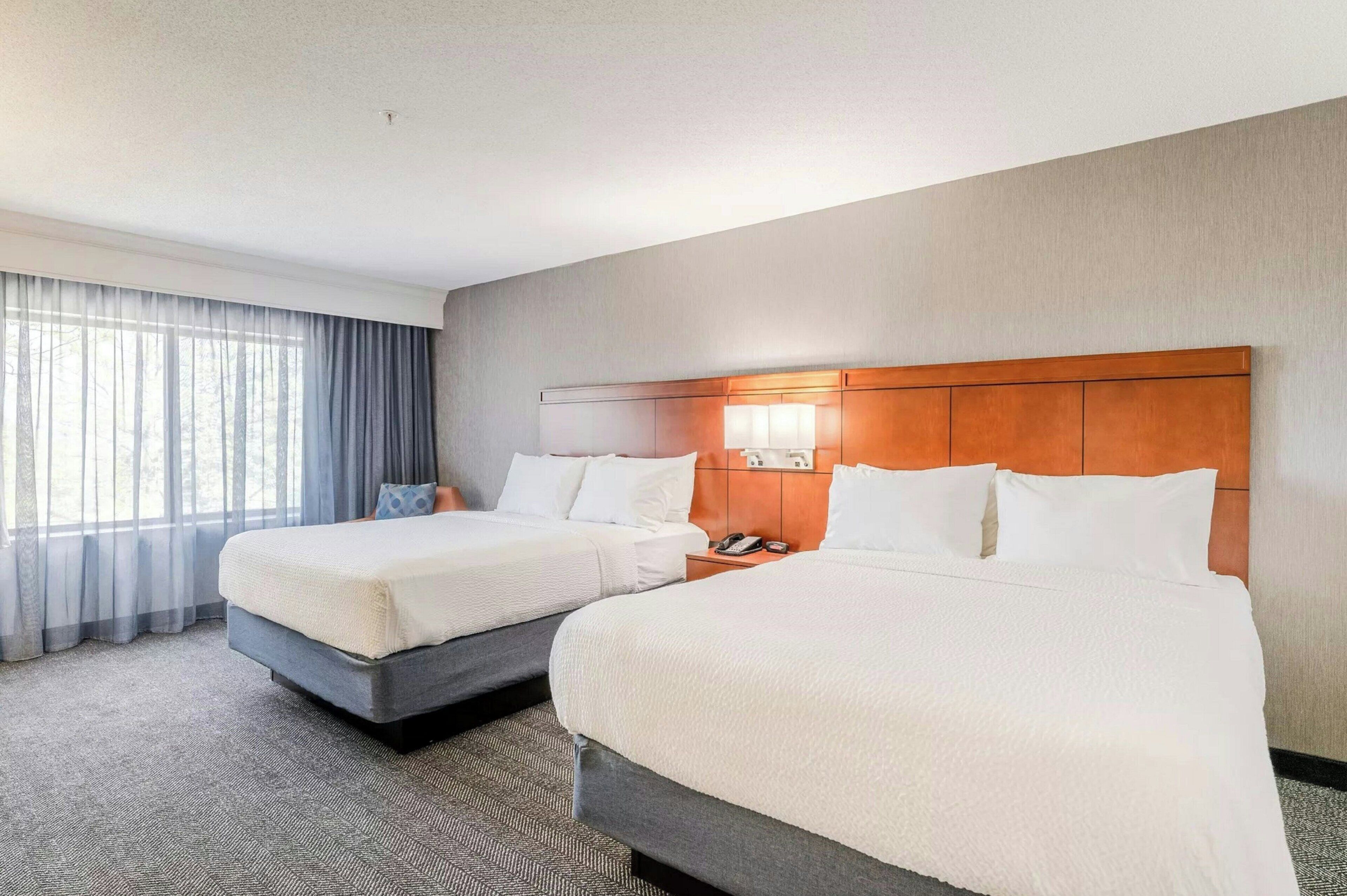 Une chambre d’hôtel avec deux lits, une tête de lit en bois, un téléphone et une fenêtre avec des rideaux.