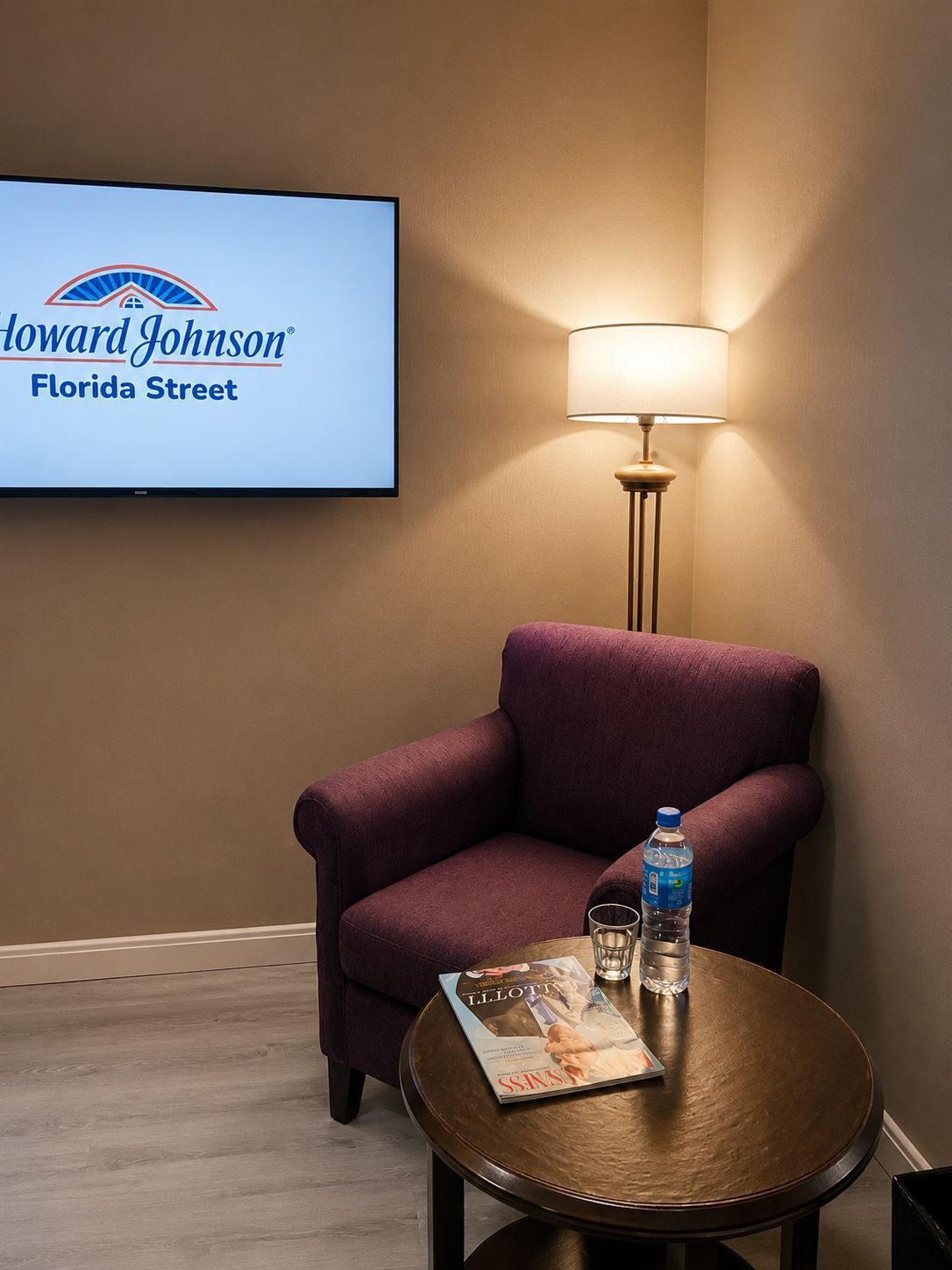 Ein Hotelzimmer mit einem violetten Sessel, einem kleinen runden Tisch mit einer Zeitschrift, einer Wasserflasche und einer Lampe. Ein Bildschirm zeigt den Hotelnamen 'Howard Johnson Florida Street'.