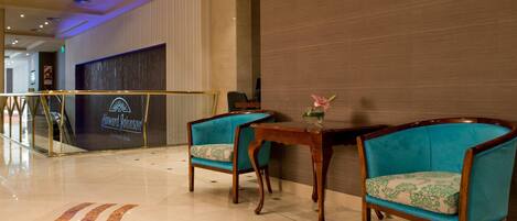 A hotel lobby with a reception desk, two blue upholstered chairs, a small wooden table, and a patterned rug.