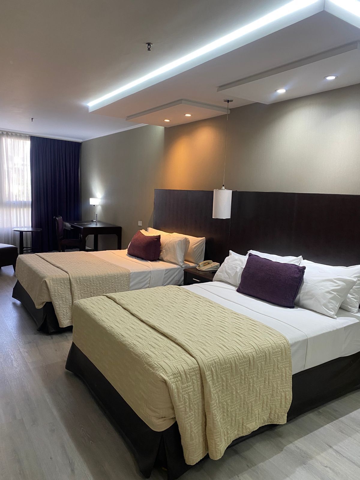 Habitación de hotel con dos camas, un escritorio, una silla, una lámpara y una ventana con cortinas.