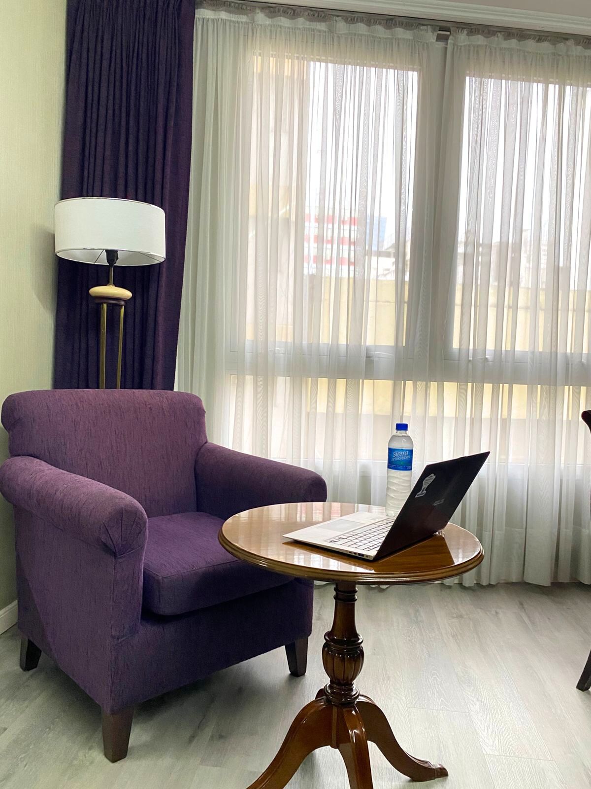 Uma poltrona roxa, uma mesa de madeira com um laptop, uma luminária e uma janela com cortinas leves.