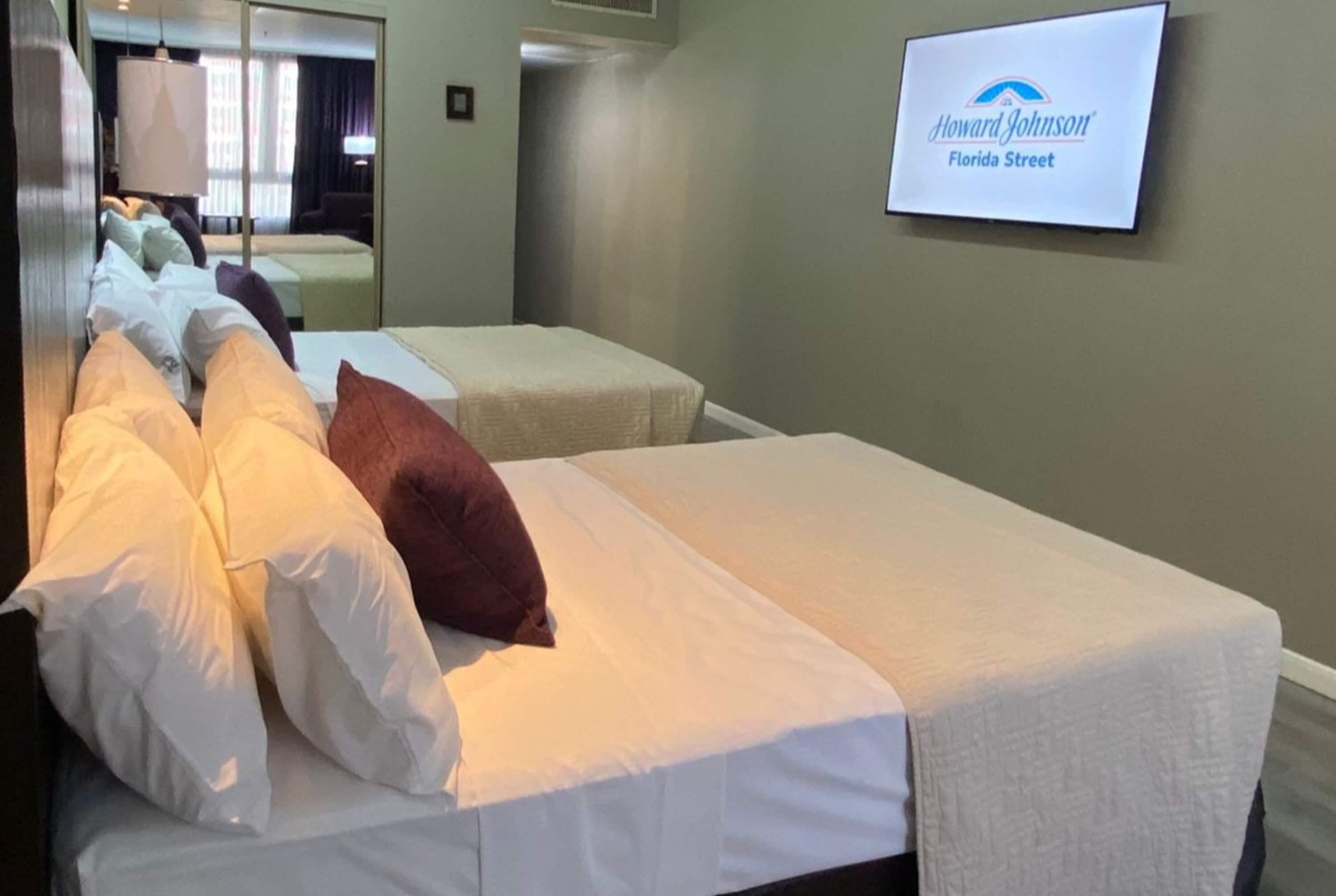 A hotel room with two beds, a television displaying the Howard Johnson logo, and a mirror reflecting the room.