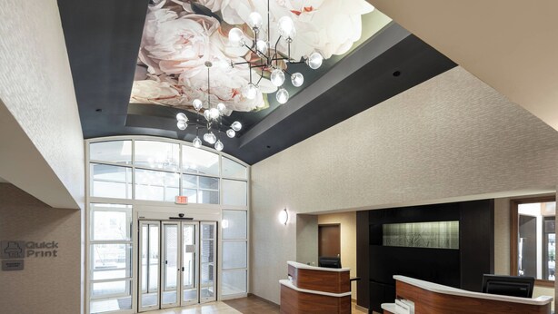 A reception area with a modern design, featuring a large glass door entrance, a wooden reception desk, and a ceiling with a floral mural. - Courtyard by Marriott Charlottesville - University Medical Center (Charlottesville)