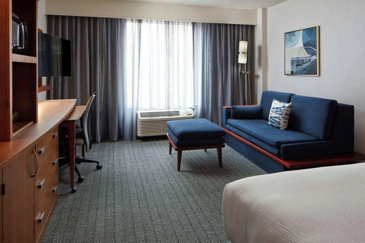 A hotel room with a desk, a blue sofa, a bed, a TV, an air conditioner, and a window with curtains.