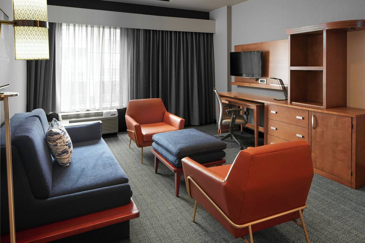 A modern hotel room with a sofa, two armchairs, a desk with a laptop, a television, and a large window with curtains.