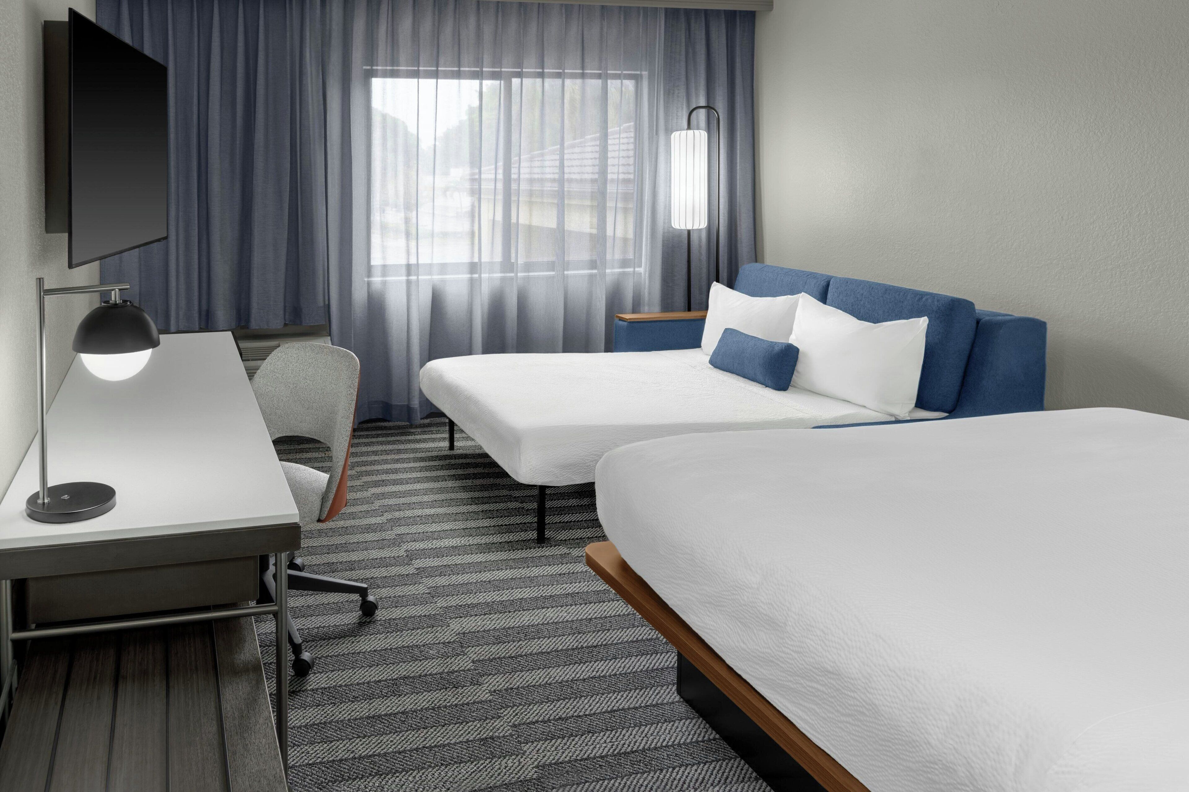 A hotel room with two beds, a desk, a chair, a TV, and a lamp.