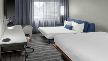 A hotel room with two beds, a desk, a chair, a TV, and a lamp.
