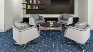 Un salón de conferencias con una mesa de cristal, cuatro sillas grises, una estantería con libros y objetos decorativos, y una gran pantalla oscura en la pared.