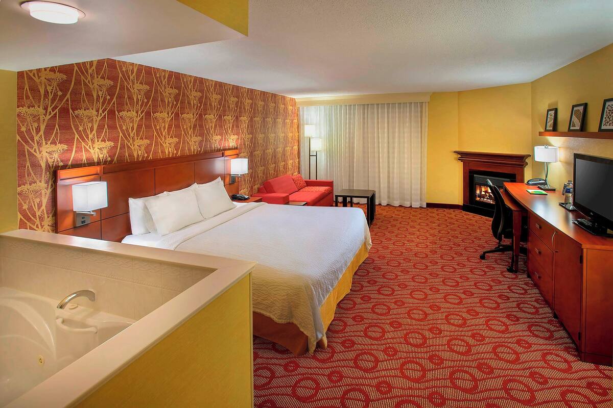 A hotel room with a large bed, a sitting area with a red sofa, a desk with a computer, a television, and a fireplace.