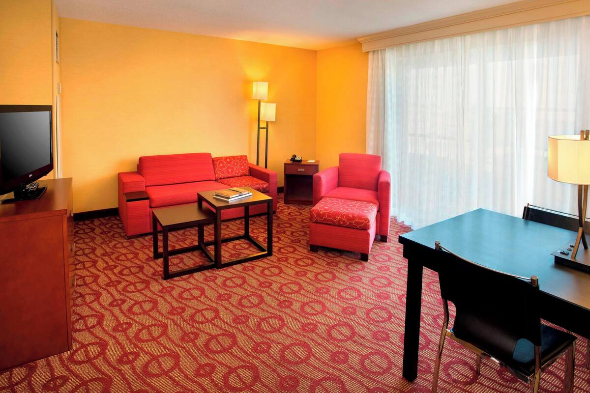 A hotel room with a red sofa, a red armchair, a small table, a TV, and a lamp.