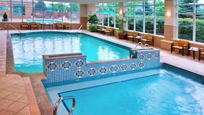 An indoor swimming pool with clear blue water, tiled edges, and a seating area with chairs and potted plants.