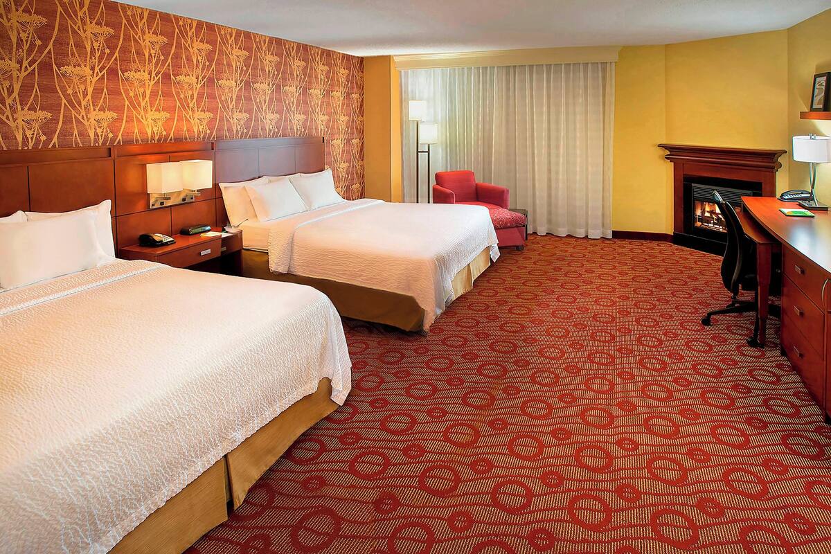 A hotel room with two beds, a red armchair, a desk with a lamp, and a fireplace.