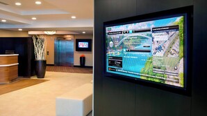 A hotel reception area with a digital screen displaying local news and weather, a reception desk, a seating area, and an elevator.