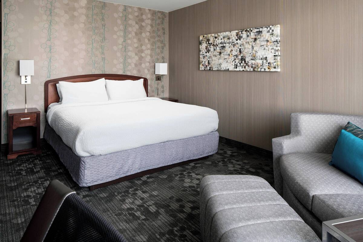 A hotel room with a large bed, a sofa, and a small table. There is a wall-mounted artwork above the bed and a nightstand with a lamp beside it.