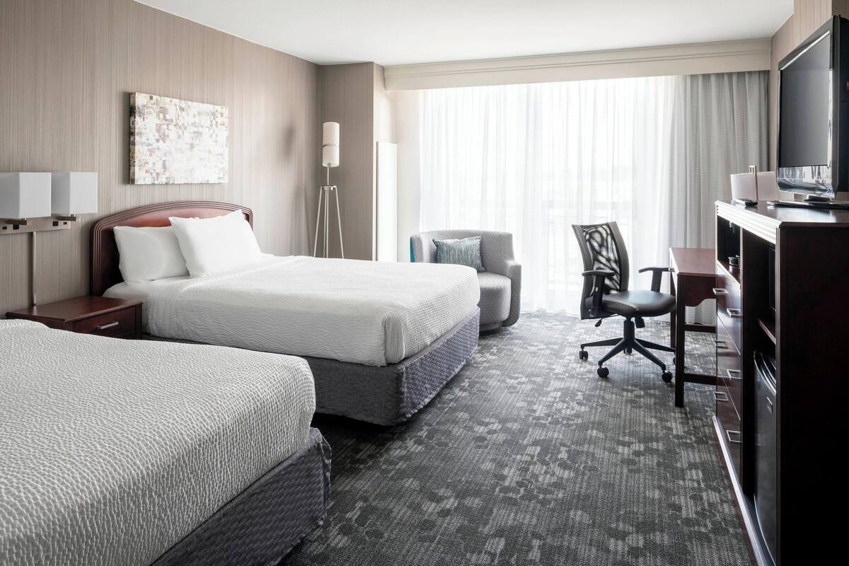 A hotel room with two beds, a desk, a chair, a TV, and a large window with curtains.