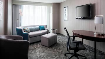 Une chambre d’hôtel équipée d’un canapé, d’un pouf, d’un bureau, d’une chaise, d’une télévision, d’une lampe et d’une grande fenêtre avec des rideaux.