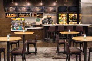 A bar area with multiple seating options, a coffee machine, and a variety of beverages and snacks displayed on shelves.