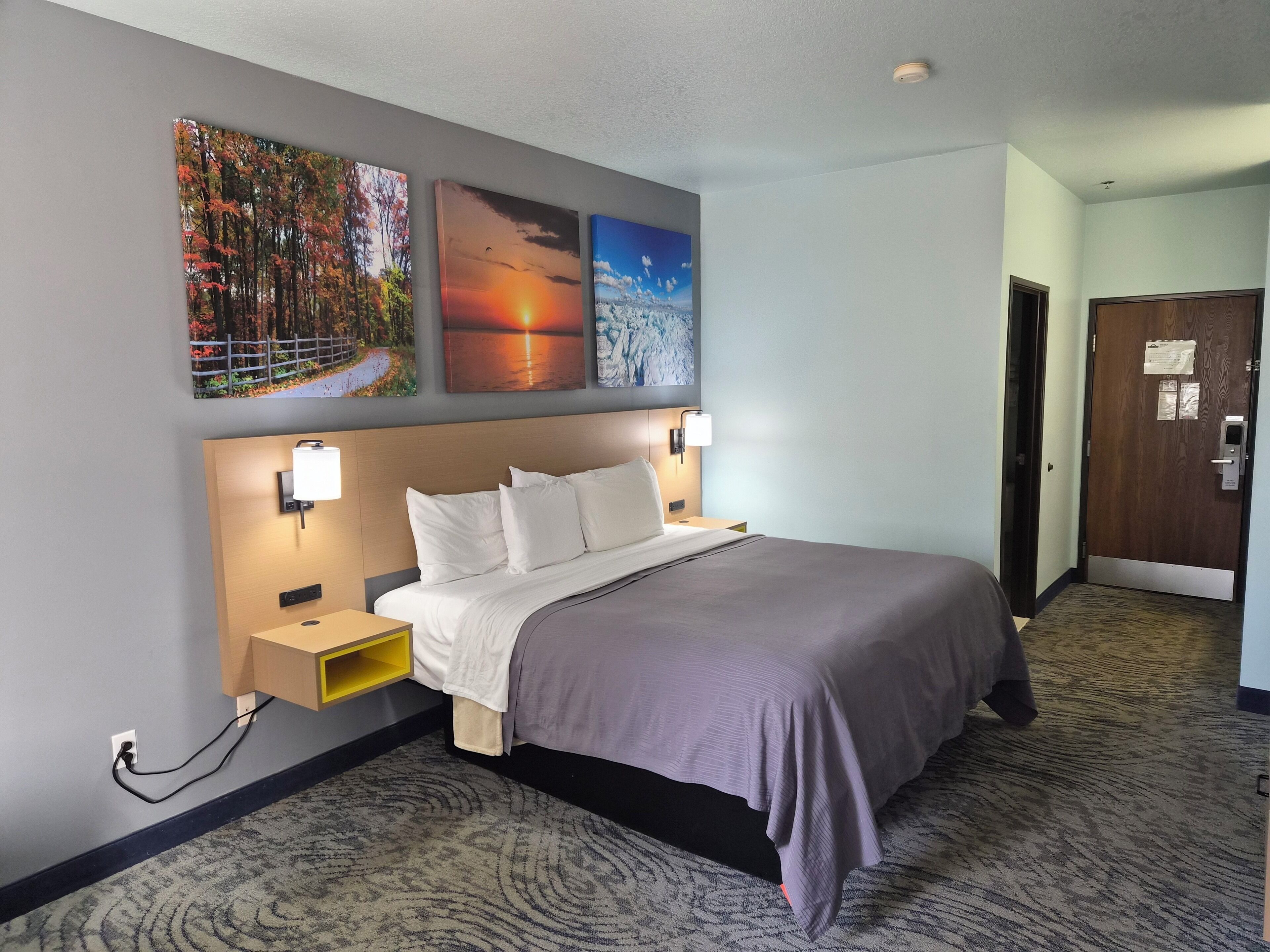 A hotel room with a bed, a nightstand, a wall-mounted lamp, and three framed pictures.