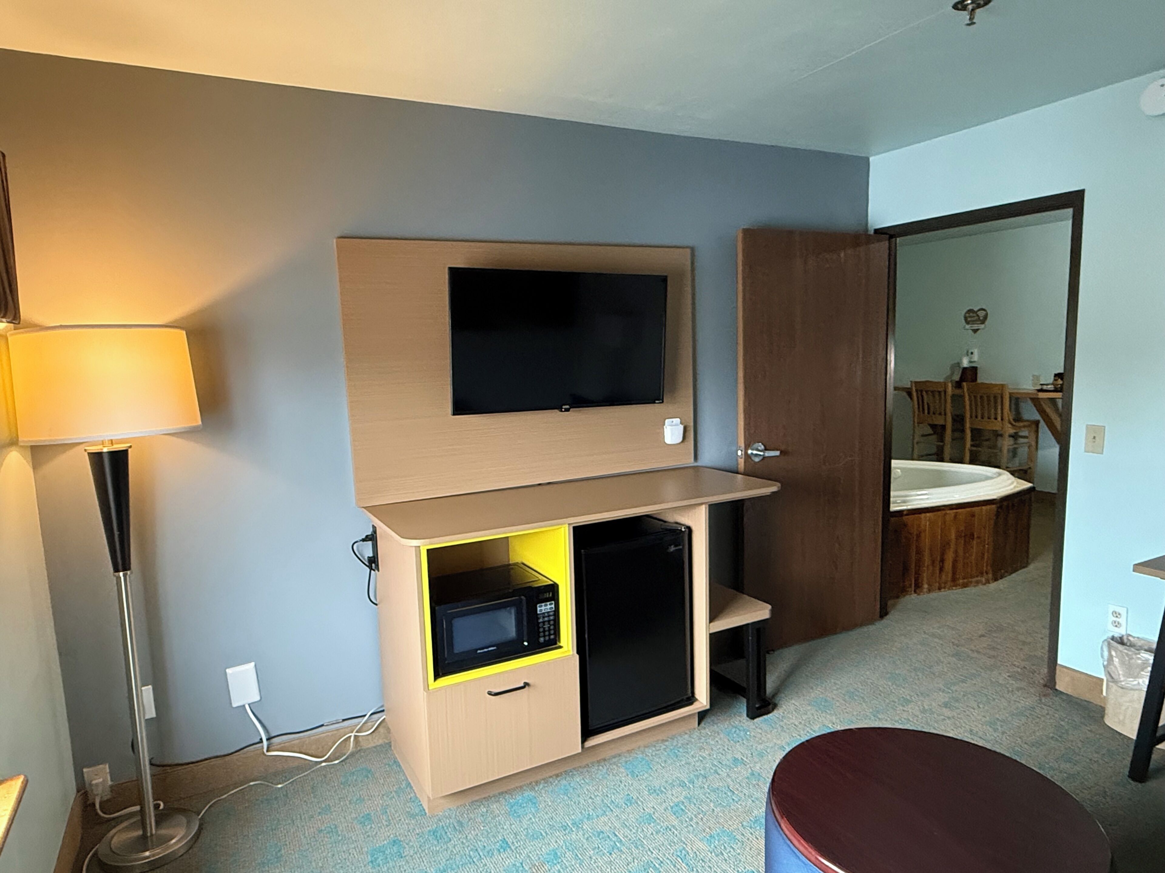 A hotel room with a TV mounted on the wall, a microwave in a cabinet, a bedside table, and a bathroom visible through an open door.