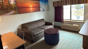 A room with a sofa, a round ottoman, a table, and a window with curtains.
