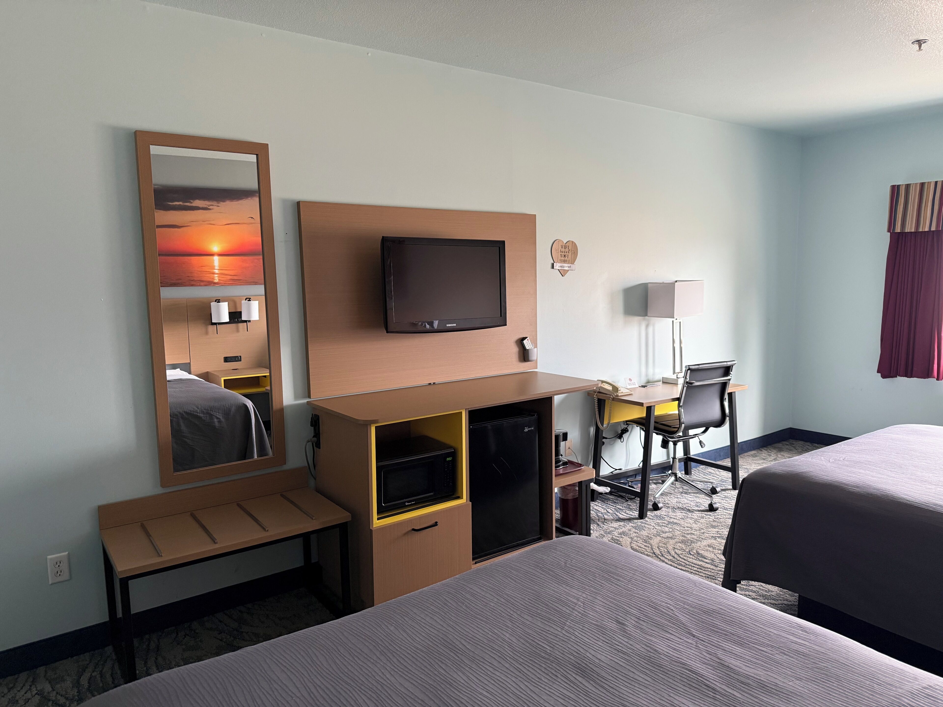 A hotel room with two beds, a desk with a chair, a television, a mirror, and a small kitchenette.