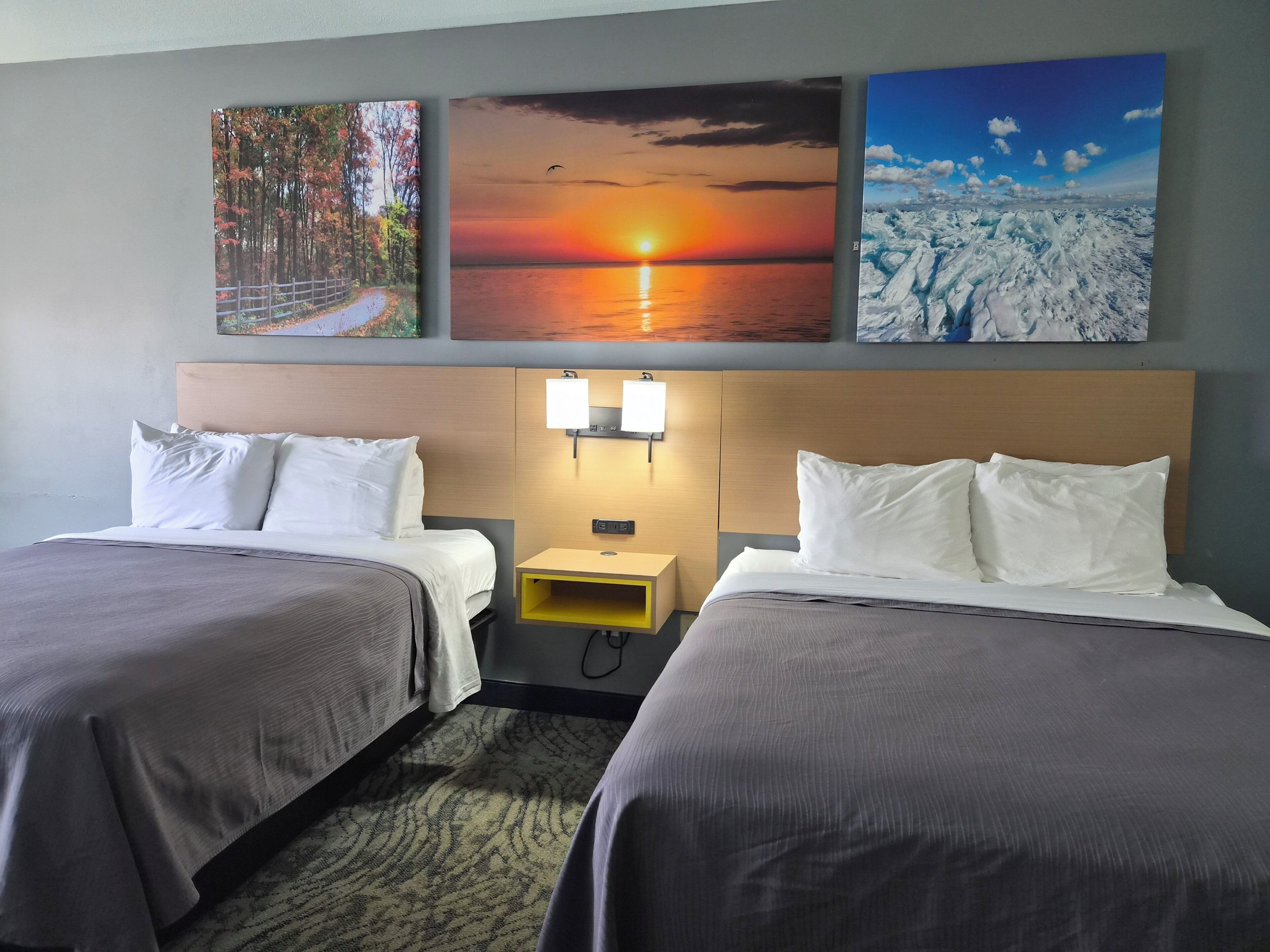 Une chambre d’hôtel avec deux lits, une tête de lit dotée de lampes fixées au mur, et trois tableaux encadrés au-dessus des lits.