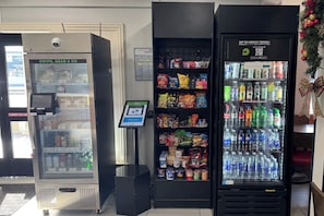A self-service kiosk with a digital screen, a refrigerated snack vending machine, and a refrigerated beverage vending machine. - Hotel 1550 - SFO Airport (San Bruno)