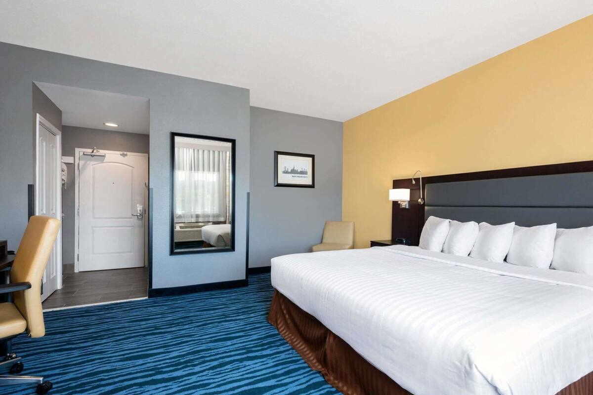 A hotel room with a large bed, a chair, a desk, a mirror, and a framed picture on the wall.