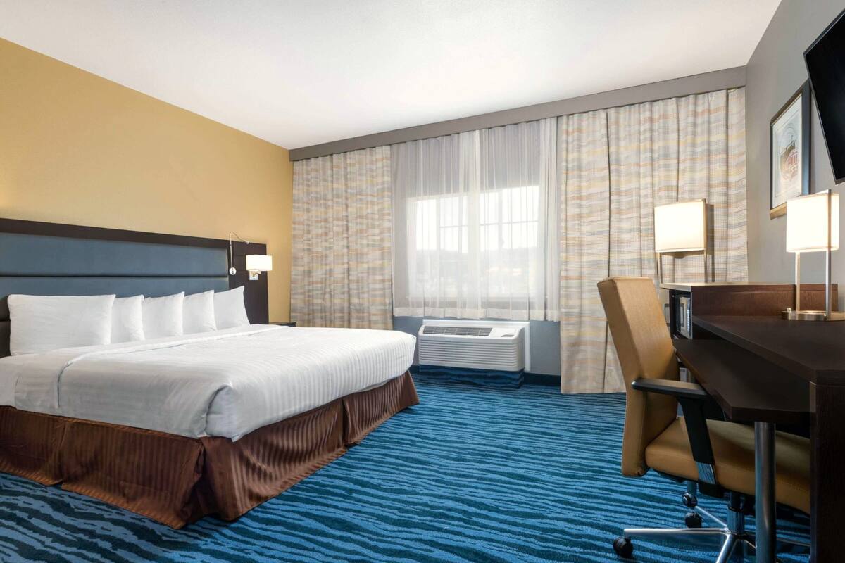 A hotel room with a bed, desk, chair, air conditioning unit, and curtains.