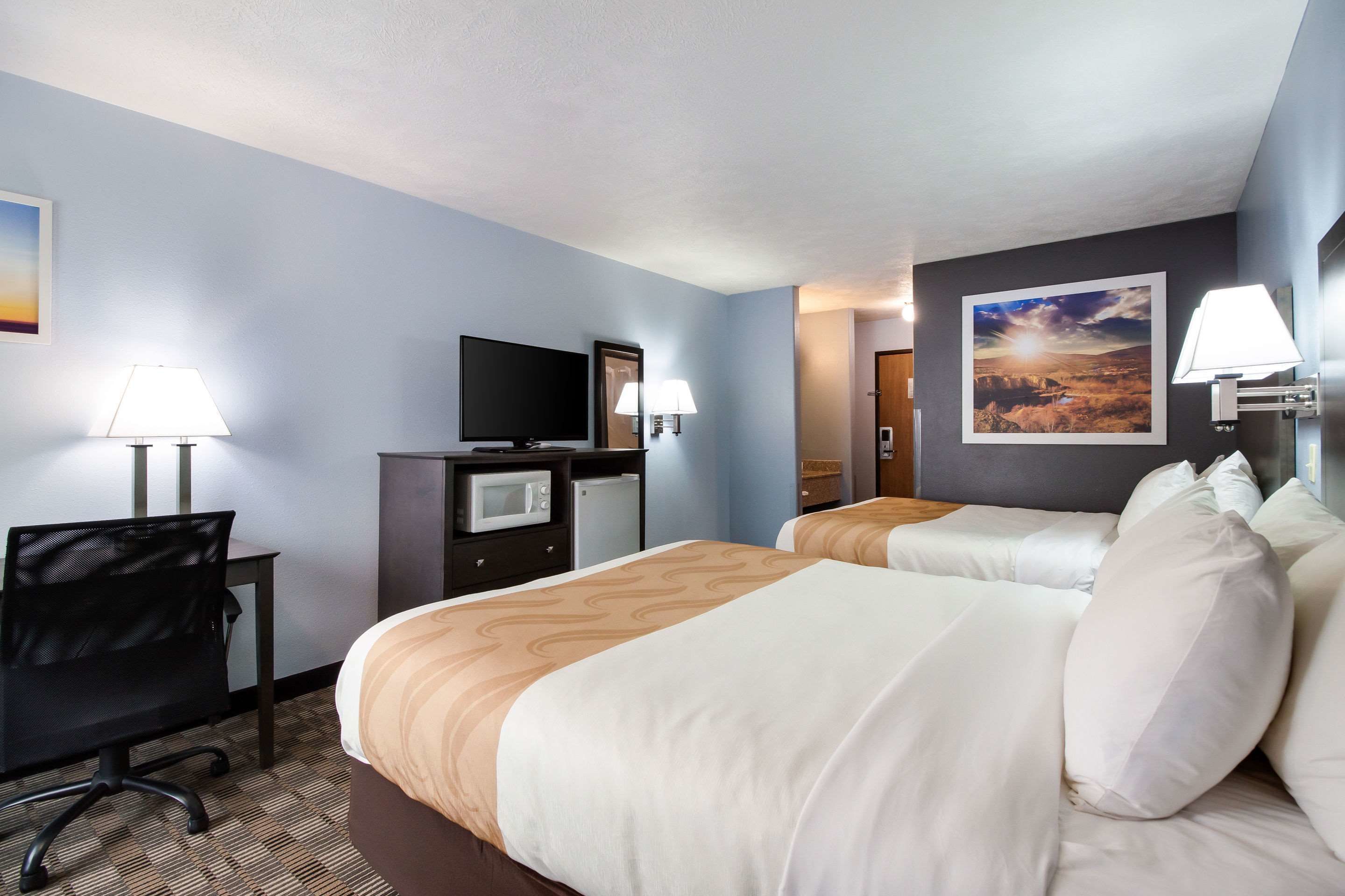 A hotel room with two beds, a desk with a chair, a television, a microwave, and a painting on the wall.