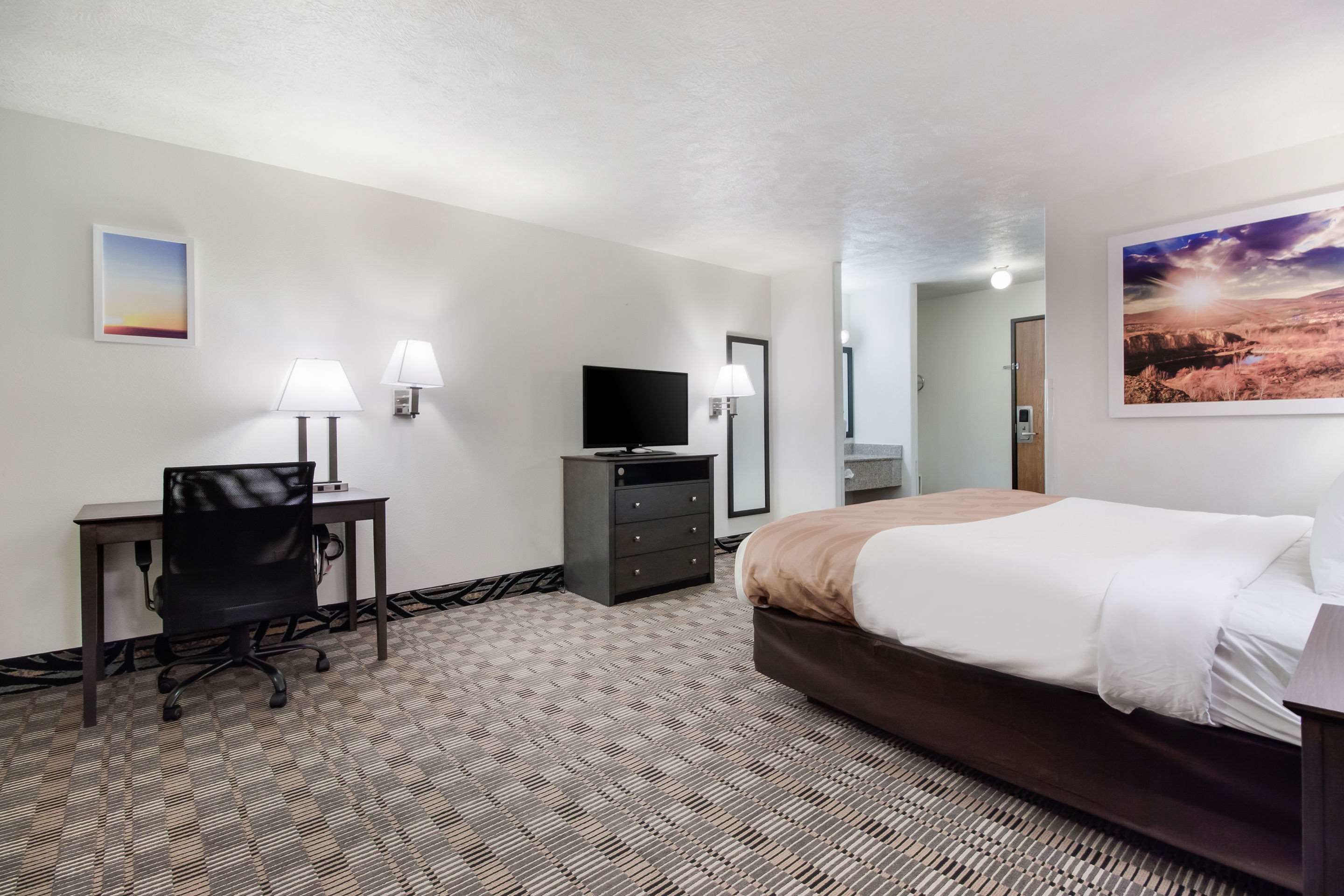 A hotel room with a bed, desk, chair, TV, dresser, and artwork on the walls.